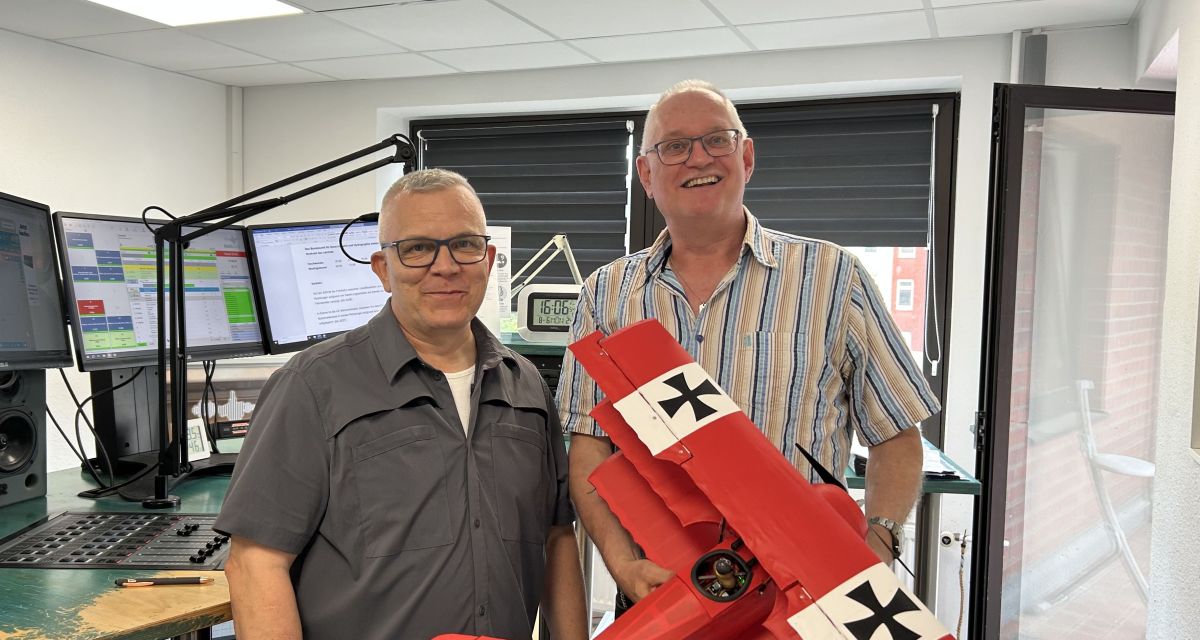 zu sehen sind zwei gutgelaunte Modellsflieger im Radiostudio, die ein etwa einen Meter großes Modell eines roten Dreideckerflugzeuges halten