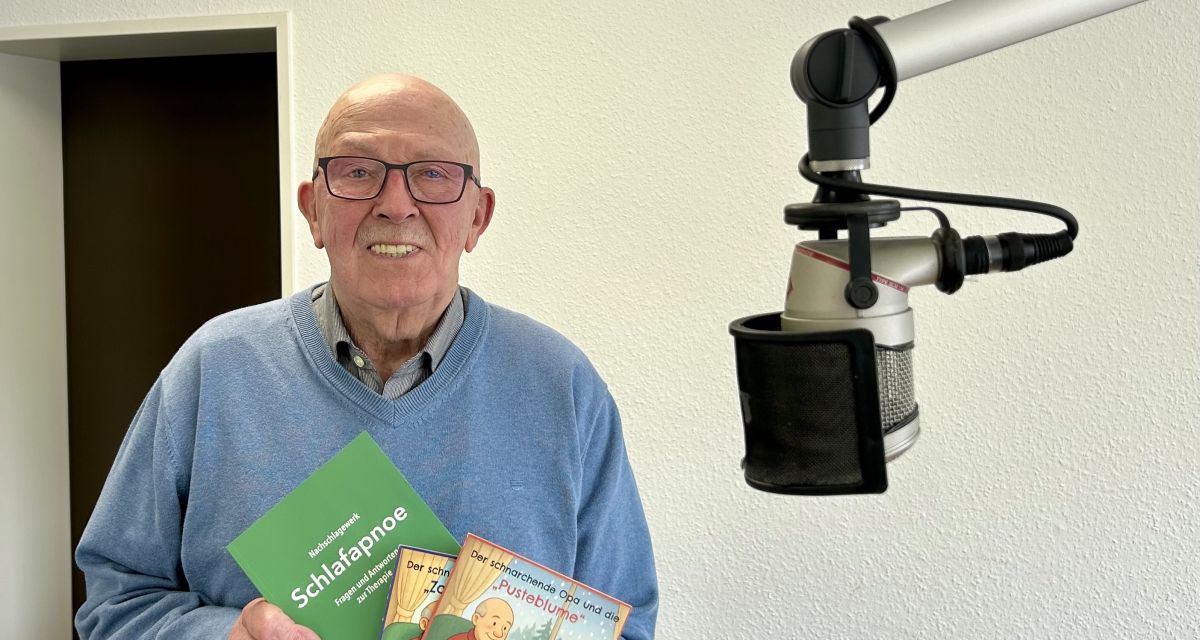 Zu sehen ist ein älterer Herr im Hörfunkstudio, der Bröschüren präsentiert.