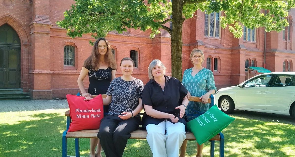 Das Konzept der Plauderbank wird am Mehrgenerationenhaus in Wilhelmshaven vorgestellt. Um eine Bank versammelt sind von links nach rechts Anke Stalling (Pastorin Banter Kirche), Mirjam Möllmann (Leiterin Mehrgenerationenhaus), Ruth Link (Initiatorin Plauderbank), Isabell Eickhoff (Senioren- und Pflegestützpunkt). Auf der Bank sind ein grünes und ein rotes Kissen mit der Aufschrift 