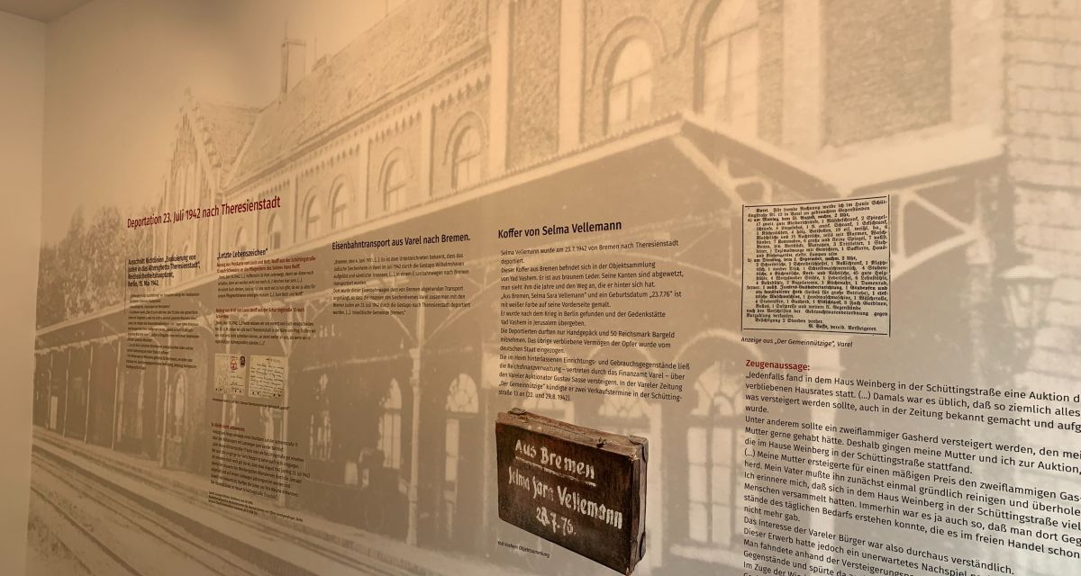 Zu sehen ist ein Teil der Ausstellung im Weinberghaus in Varel. Die AUsstellung behandelt die jüdische Geschichte in der Region. Auf dem Bild zu sehen ist eine mit dem Bild eines Bahnhofes bedruckte Wand, darauf ist ein Informationstext zur Überschrift 