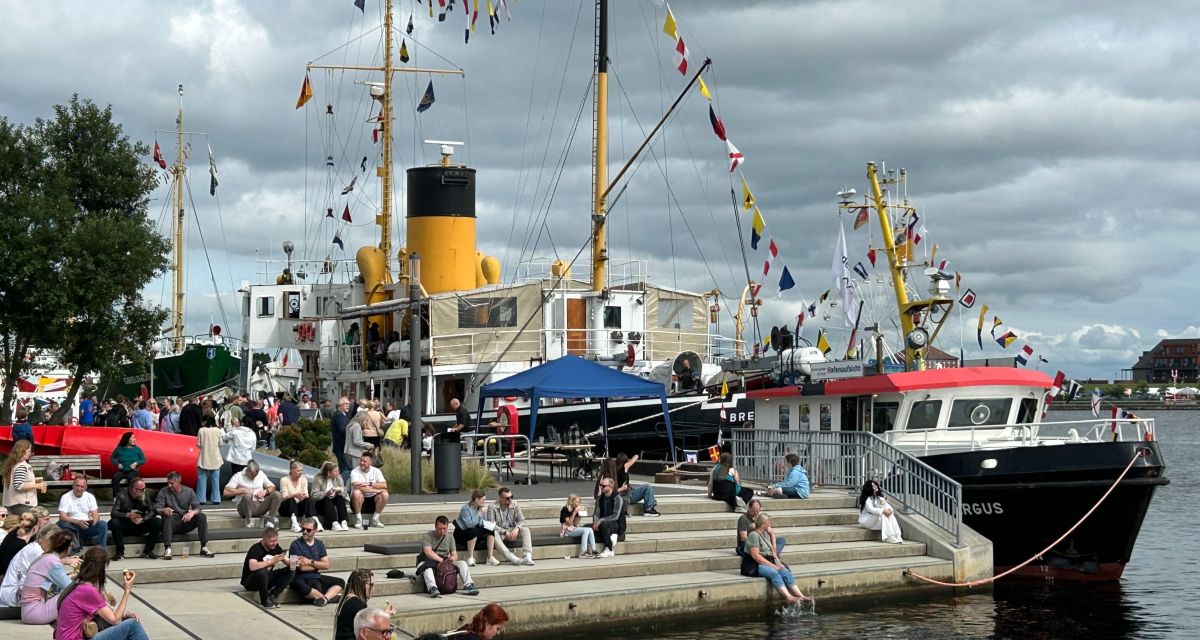 Über die Toppen geflaggte Schiffe an den Terrassen des Museumshavens, die zum Stadtfest gut bevölkert sind. Der Himmel ist wolkig.