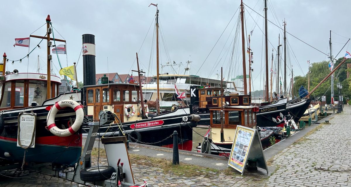 Zu sehen ist der Museumshafen in Leer. Alte Schiffe liegen an einer Kaimauer, am Rand ist ein gepflasterter Fußweg zu sehen.