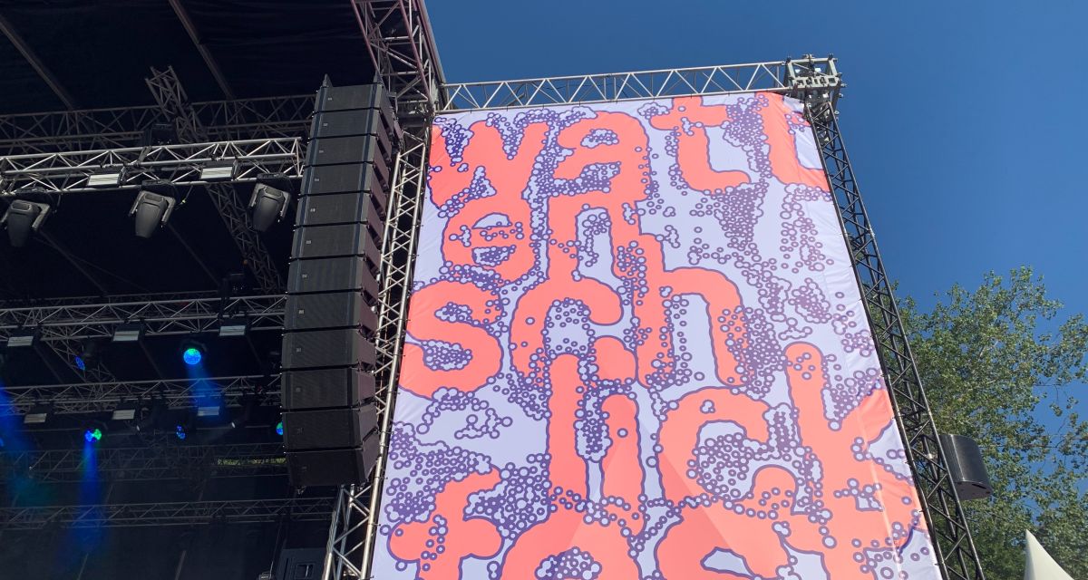 Auf dem Foto ist eine große Leinwand neben einer Festivalbühne zu sehen. In rosa und lila-blauer Schrift steht dort
