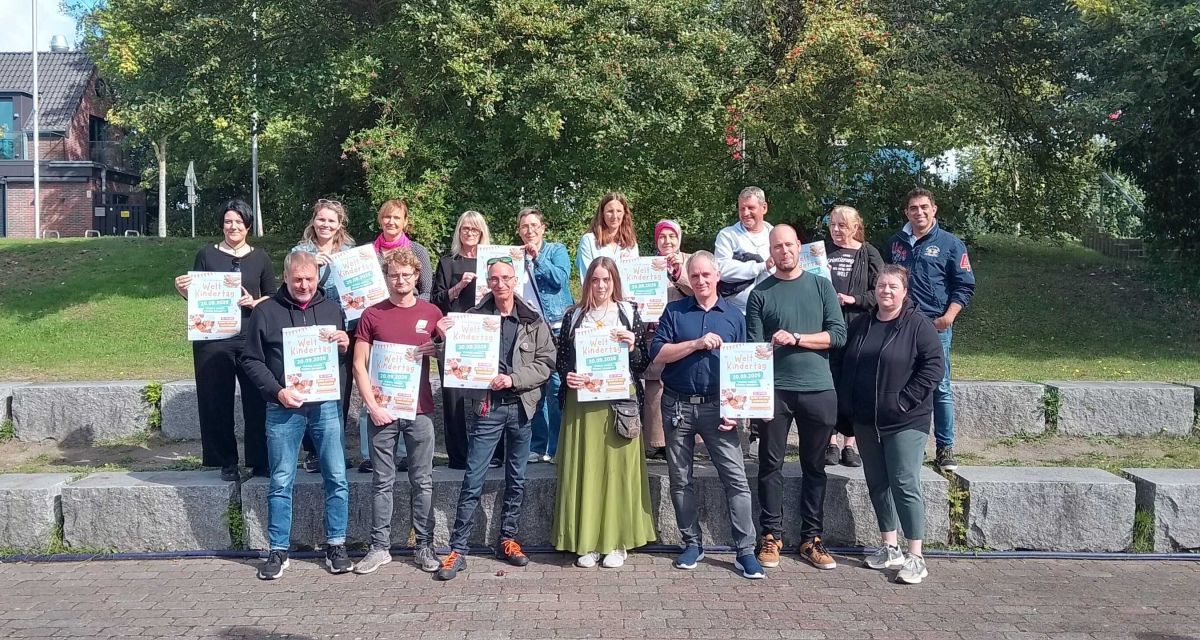 Organisator*innen und Mitwirkende des Kinderfests in Wilhelmshaven für 2026 stehen auf der Steintreppe im Pumpwerkpark und halten das Plakat der Veranstaltung in den Händen.