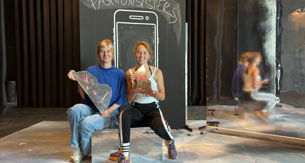 Regisseurin Güde Nissen und Schauspielerin Nora Kelschebach in der Kulisse zu Heimatkleid. Beide tragen Streetwear und halten Tafelstücke mit Gesichtern in den Händen. Im Hintergerund steht auf einer Tafel 