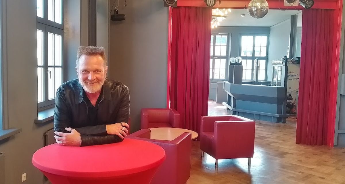 Der Intendant der Landesbühne Niedersachsen Nord, Olaf Strieb, im Oberen Foyer des Stadttheaters Wilhelmshaven. Er steht an einem mit rotem Tuch bespannten Stehtisch und hat sich aufgestützt. Der lange Raum mit Parkett ist außerdem mit roten Sitzmöbeln bestückt.