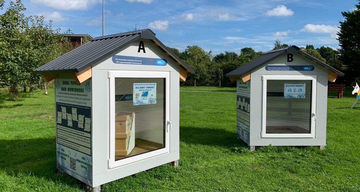 Zu sehen sind die zwei Hütten, die für die Eisblockwette gebaut wurden. Beide sind komplett baugleich, aber eine ist gedämmt, die andere nicht. Die kleinen Hütten sind grau udn haben vorne eine Fensterscheibe installiert. Darin werden 100 Liter gefrorenes Wasser aufbewahrt, und es kann gewettet werden, wie groß die Differenz im Schmelzwasser ist. Die Eisblockwette findet vom 22.09.26 bis zum 27.09.26 im Störtebeker-Park in Wilhelmshaven statt. 