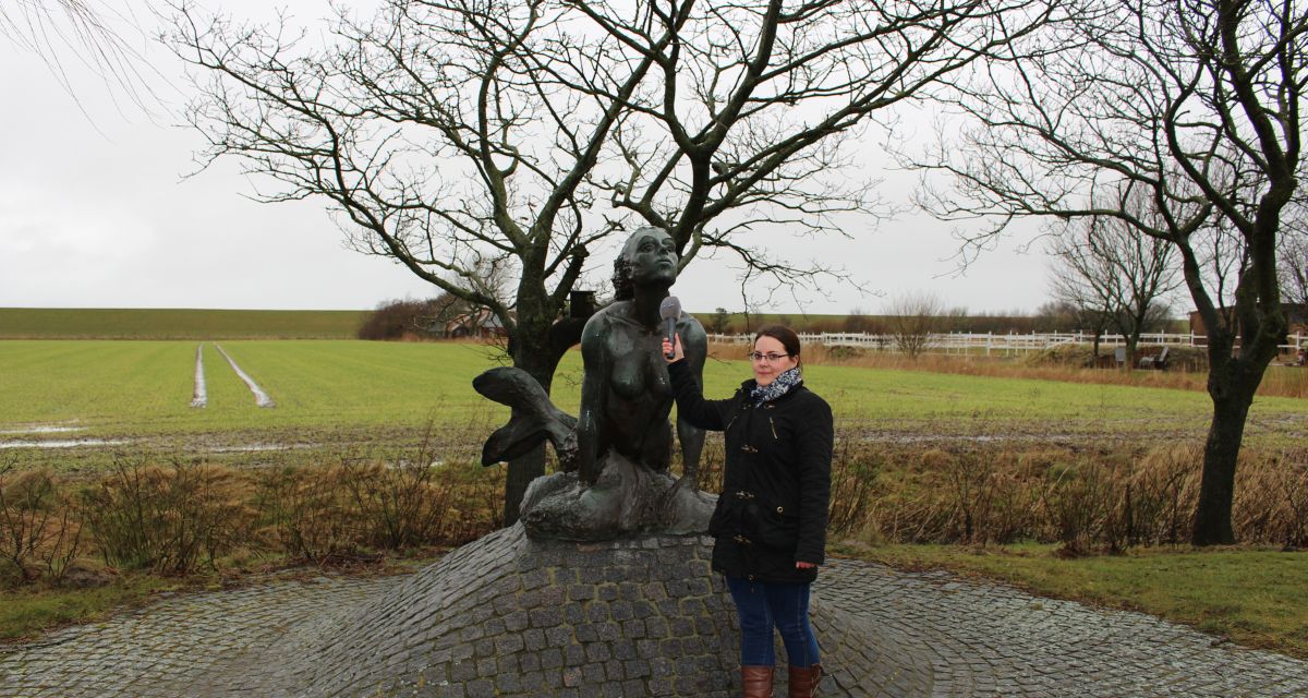 Eine junge Frau mit Mikrophon tut so, als ob sie das Denkmal einer Meerjungfrau interviewe.
