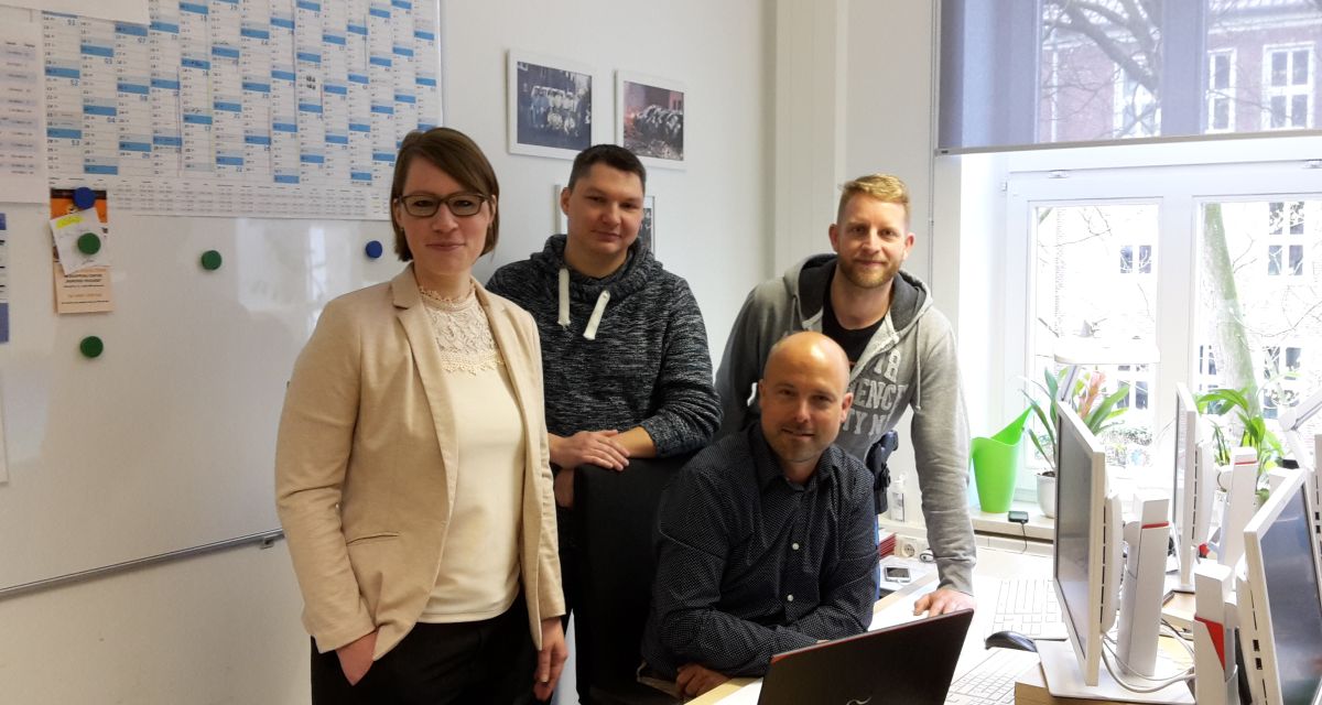 Man sieht eine Frau und drei Männer im Büro für die Kamera posieren.