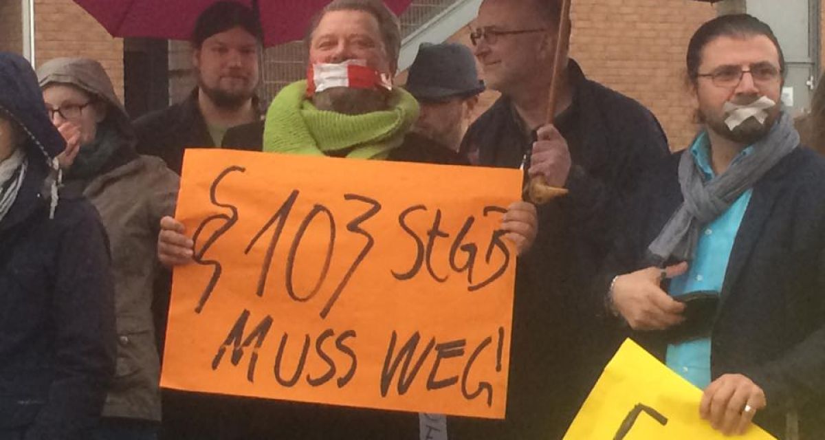Menschen mit zugeklebten Mündern halten ein Schild hoch, auf dem steht: Paragraph 103 StgB muss weg.