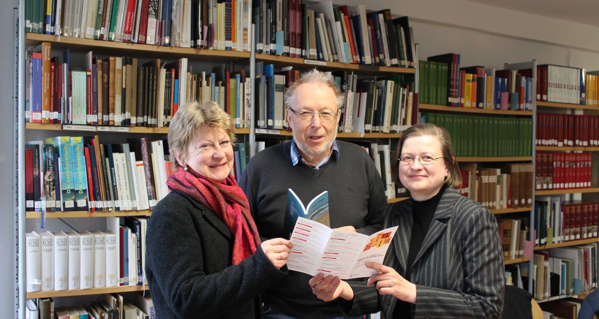 Drei Menschen lesen vor einer Bücherwand einen Flyer.