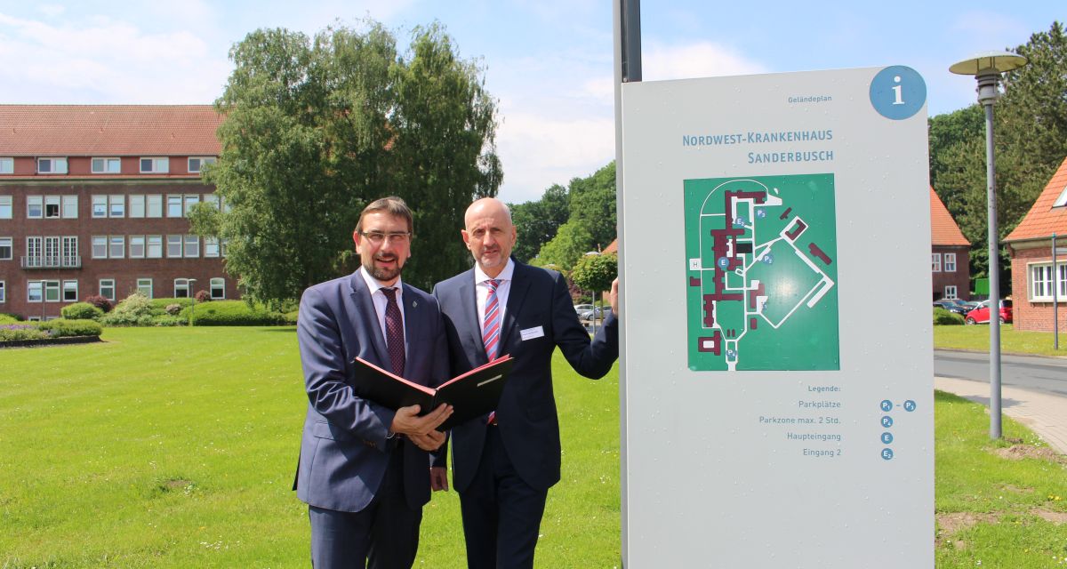 Zwei Männer posieren vor einem Lageplan-Schild des Nordwest-Krankenhauses in Sanderbusch.