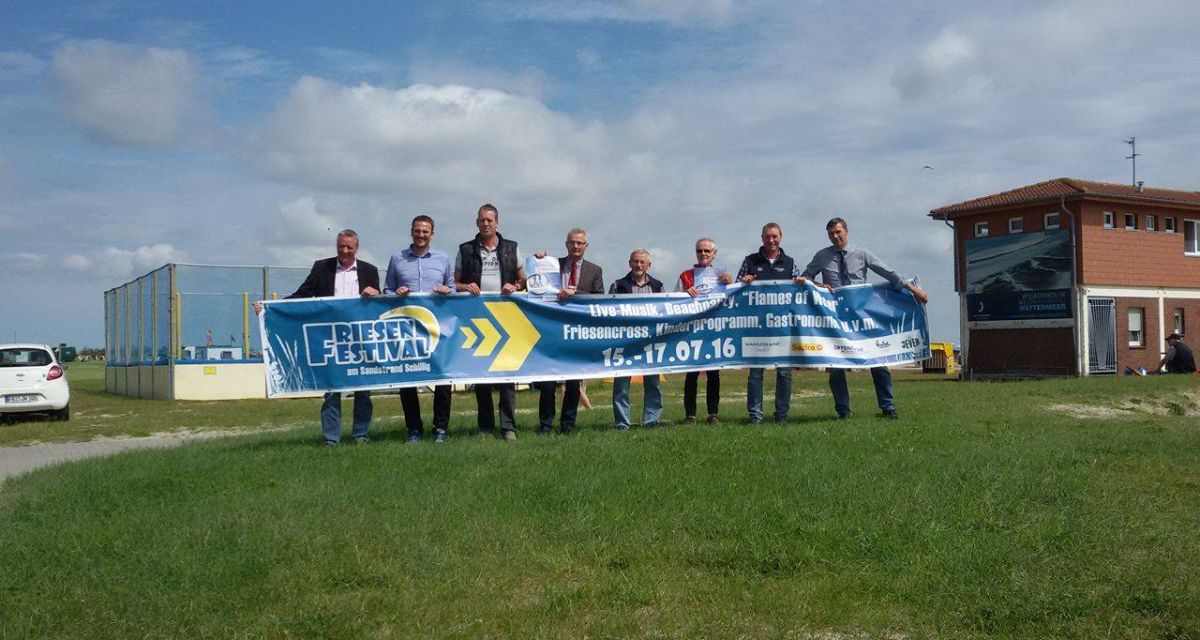 Auf grüner Wiese halten mehrere Menschen vor leicht bewölktem Himmel das Banner fürs Friesen Festival.