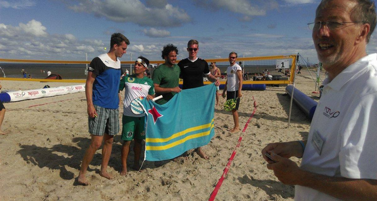 Das WHV-Team und das Team aus Aruba stehen auf dem Feld auf dem Sand und halten eine blaue Fahne mit zwei gelben Streifen in den Händen. 