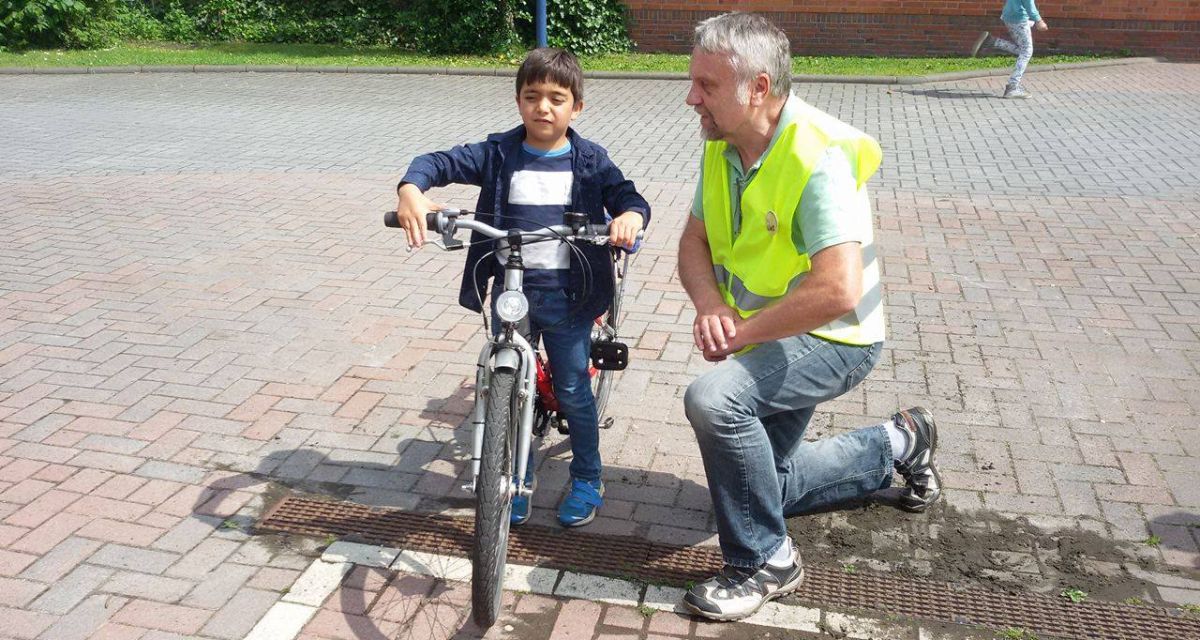 Ein Mann mit Sicherheitsweste kniet neben einem Jungen auf einem Fahrrad.
