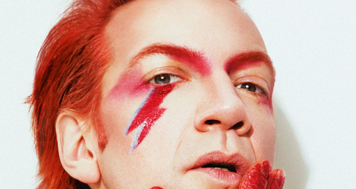 Zu sehen ist ein auffällig geschminkter, rothaariger Mann. Er hat zudem rot gefärbte Augenbrauen und Fingerspitzen, dazu trägt er einen roten Blitz unter dem rechten Auge.