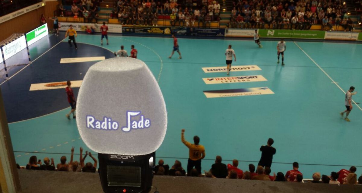 Auf dem Bild ist im Vordergrund ein Radio Jade Mikrophon zu sehen. Im Hintergrund das laufende Handballspiel.