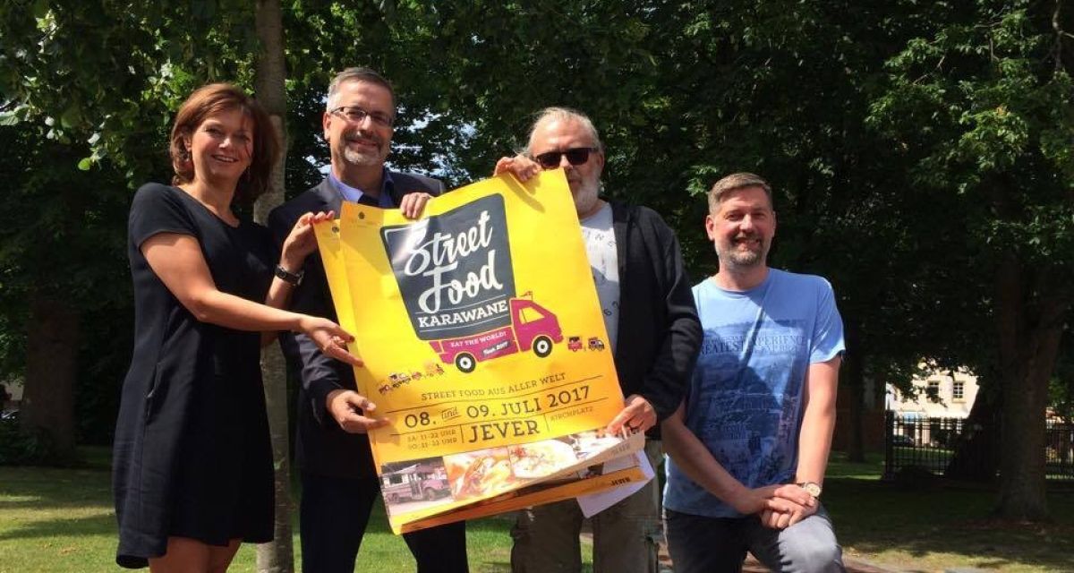 Vier Personen zeigen ein Plakat der Street Food Karawane in Jever in die Kamera. 