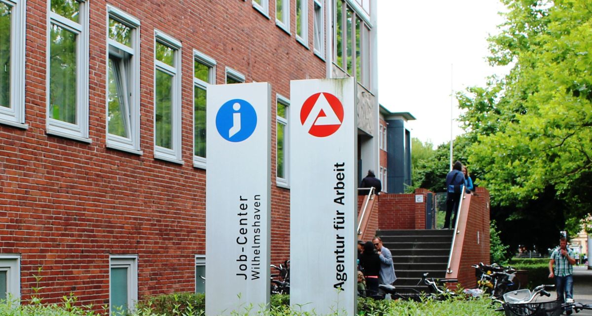 Die Agentur für Arbeit Oldenburg Wilhelmshaven mit ihrem Standort in der Jadestadt. Vor dem roten Klinkergebäude sind die Schilder der Agentur und des Jobcenters zu sehen. Dahinter liegt der Treppenaufstieg zum Eingangsbereich.