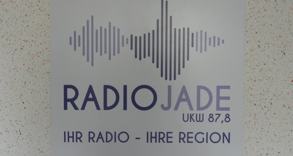 Ein Schild mit Radio Jade Logo ist an einer weißen Wand mit bunten Punkten befestigt.