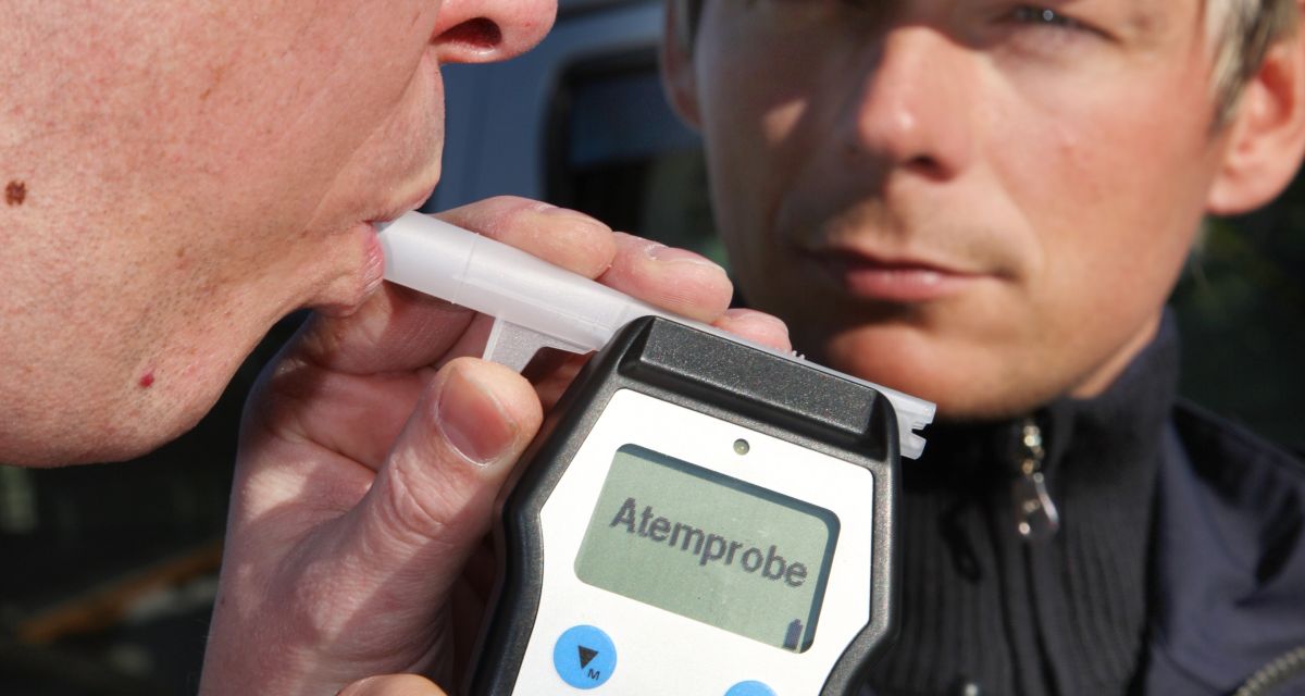 Ein Mann pustet in ein Atemalkohol-Messgerät, welches ein Polizist hält. 