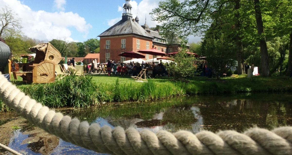 Das Schloss Gödens im Sonnenschein. Davor ein Wiese mit vielen Menschen. 