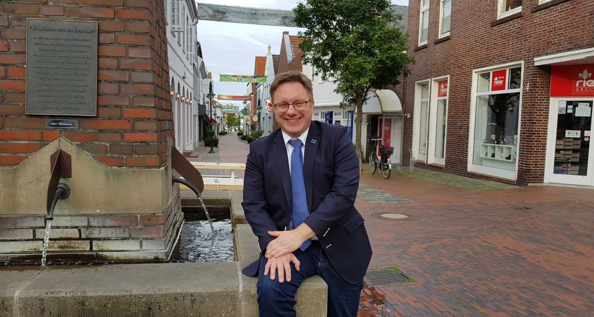 Jever Bürgermeister Jan Edo Albers sitzt an einem Brunnen in der Stadt und lacht. 