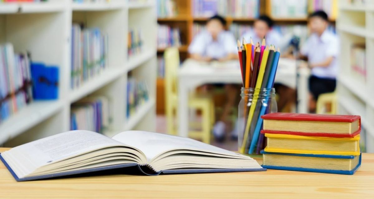 Zu sehen sind Bücher und Stifte vor einem Regal und drei Schülern.