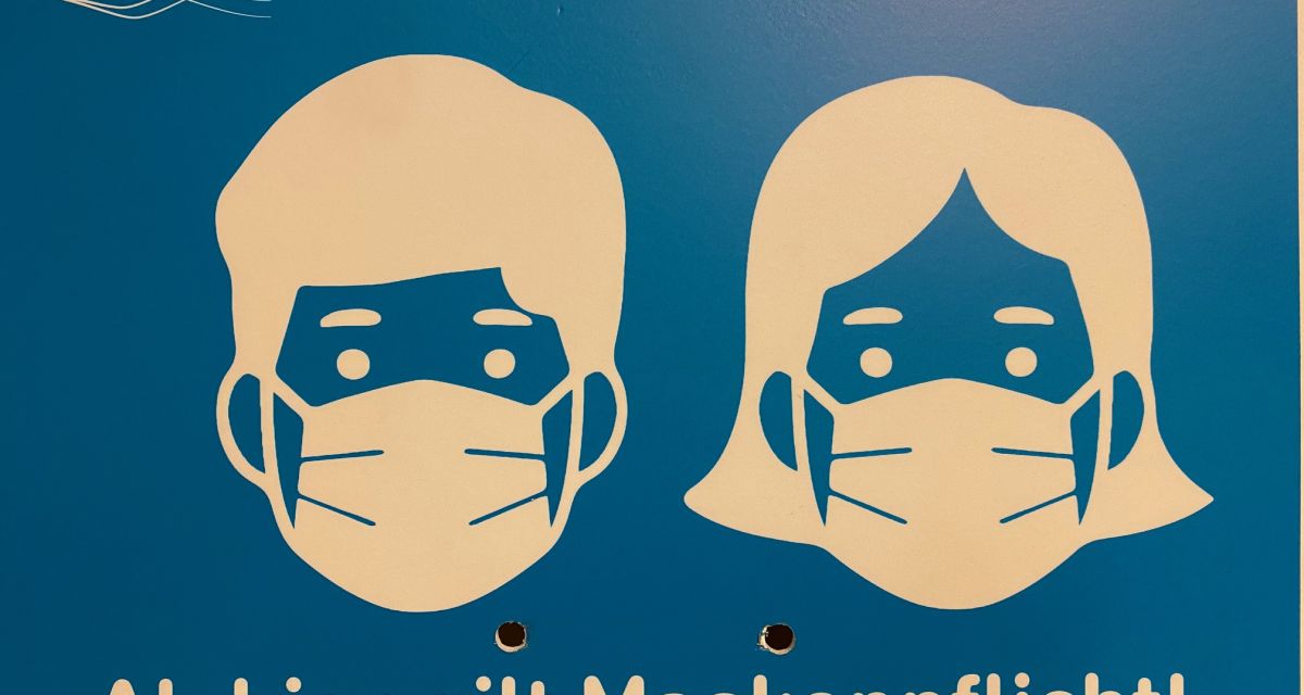 Zu sehen ist ein Schild der Stadt Wilhelmshaven auf dem mithilfe der Abbildung von zwei Menschen mit medizinischen Masken und der Aufschrift 