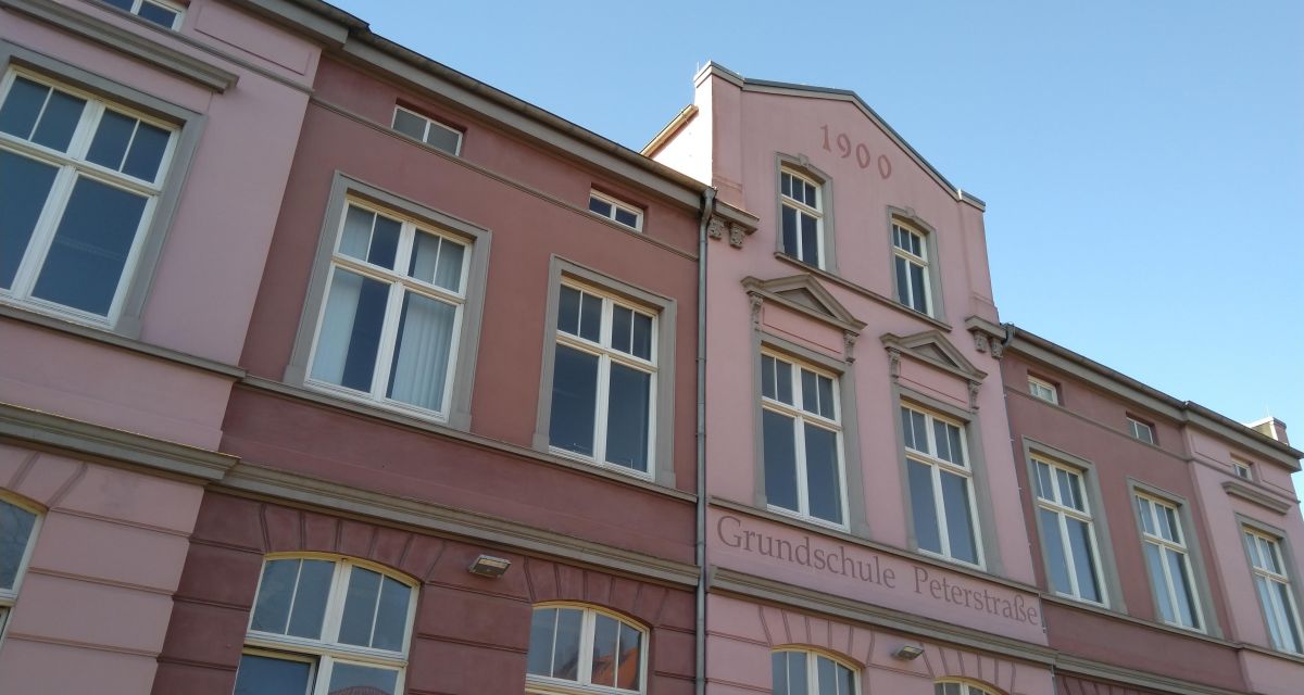 Zu sehen ist die Stirnseite des Hauptgebäudes der ehemaligen Grundschule Peterstraße in Wilhelmshaven. Das Gebäude ist in Rosatönen gestrichen, an der Fassade stehen die Worte 