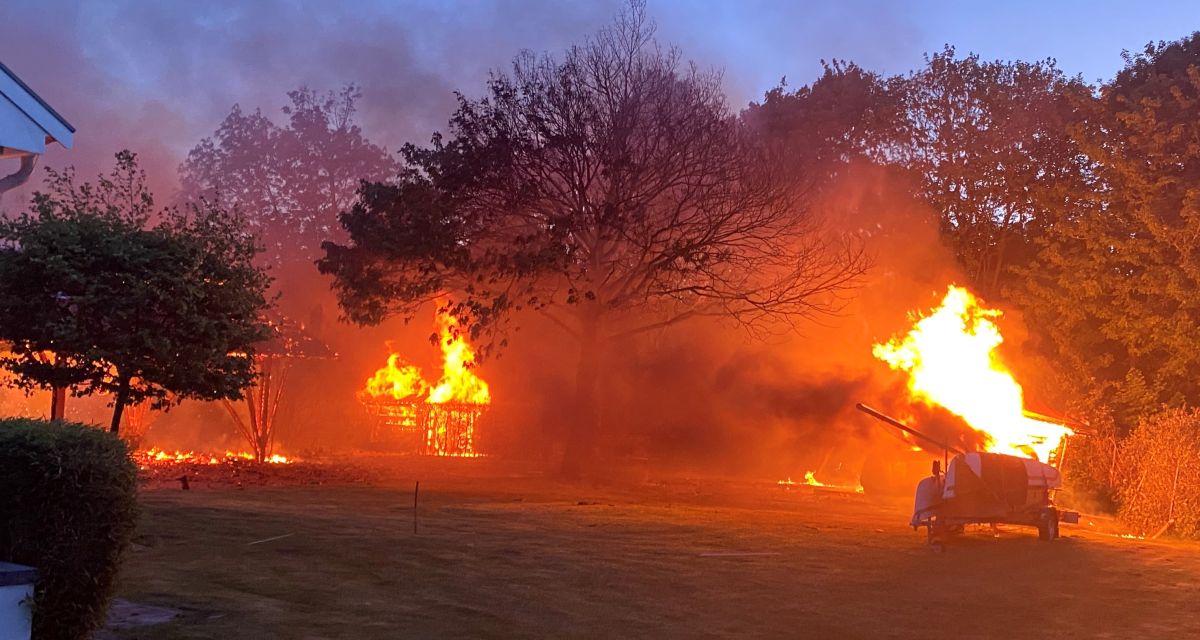 Zu sehen sind brennende Schuppen nach der Flüssiggastank Explosion. Der schwarze Rauch zieht über den Blauen Himmel. Im Hintergrund sind viele Bäume zu sehen die zur frühen Morgenzeit nur wenig durch die Sonne beleuchtet sind.