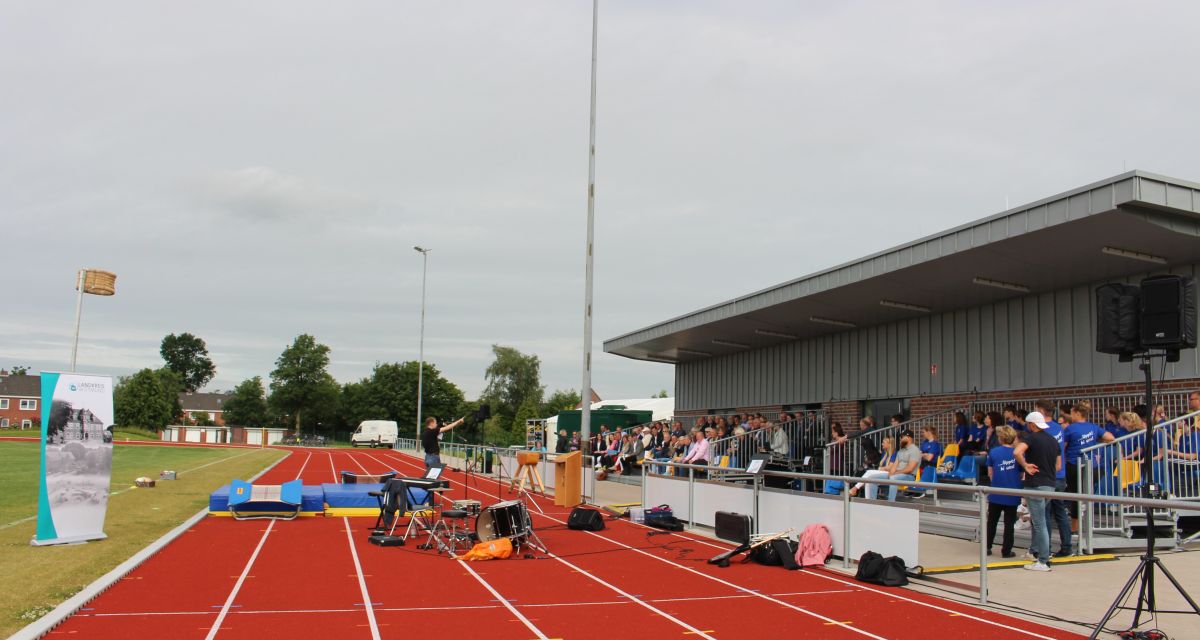 Das frisch sanierte Stadion in Wittmund an der Breslauer Straße wird festlich wiedereröffnet. Vertreter*innen von Kreis und Stadt, Sport und Kultur sitzen auf der Zuschauer*innentribüne, auf der Laufbahn steht ein Mann inmitten von Musikinstrumenten. Link im Bild ist ein Banner des Landkreises Wittmund zu erkennen.
