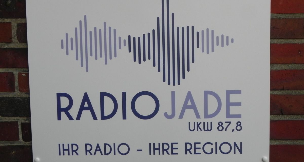 Ein Schild mit Radio Jade Logo hängt an einer Klinkerwand.
