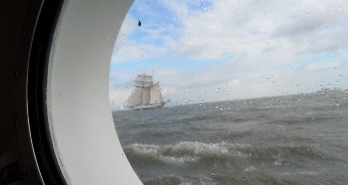 Aus einem Bullauge heraus sieht man das Meer und ein Segelschiff 