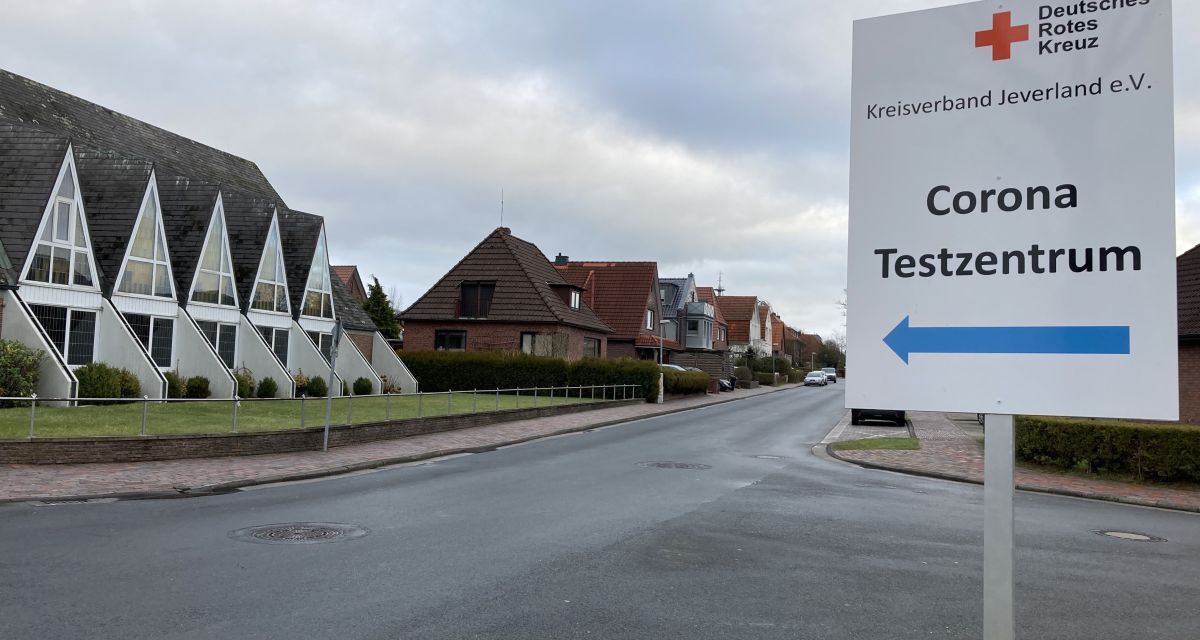 Auf dem Bild ist ein Schild mit Pfeil nach links zu sehen. Es zeigt den Weg zum Corona-Testzentrum des DRK Jever. Das Schild steht am Bahnhof. Die Straße dahinter ist komplett leer. 