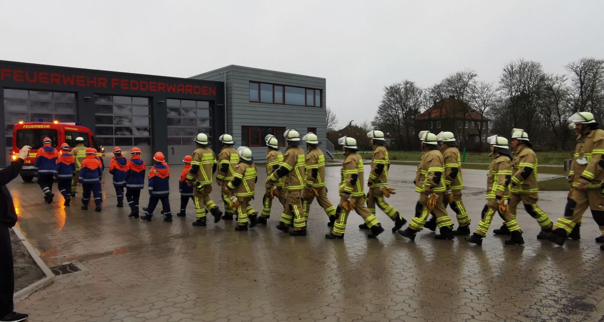 Kräfte der Freiwilligen Feuerwehr Fedderwarden sind bei regnerischem Wetter in Einsatzmontur auf dem Weg in ihr neues Feuerwehrhaus. Sie gehen in Zweierreihen nacheinander von rechts nach links durchs Bild.