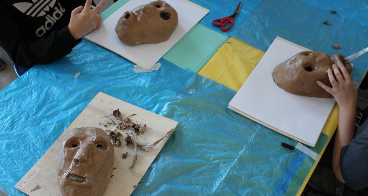 Ein Bild aus einem Workshop der 11. Theaterfabrik an der Grundschule Rheinstraße in Wilhelmshaven. Auf einem mit blauen Müllsäcken ausgelegten Tisch liegen drei Masken, die im Workshop entstehen. Vom Bildrand sind die Hände zweier Kinder bei der Arbeit zu sehen.