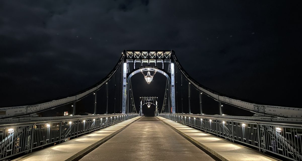 Auf dem Bild ist die Kaiser-Wilhelm-Brücke bei Nacht zu sehen. Es zeigt zentriert den Torbogen der Drehbrücke und die Fahrbahn.