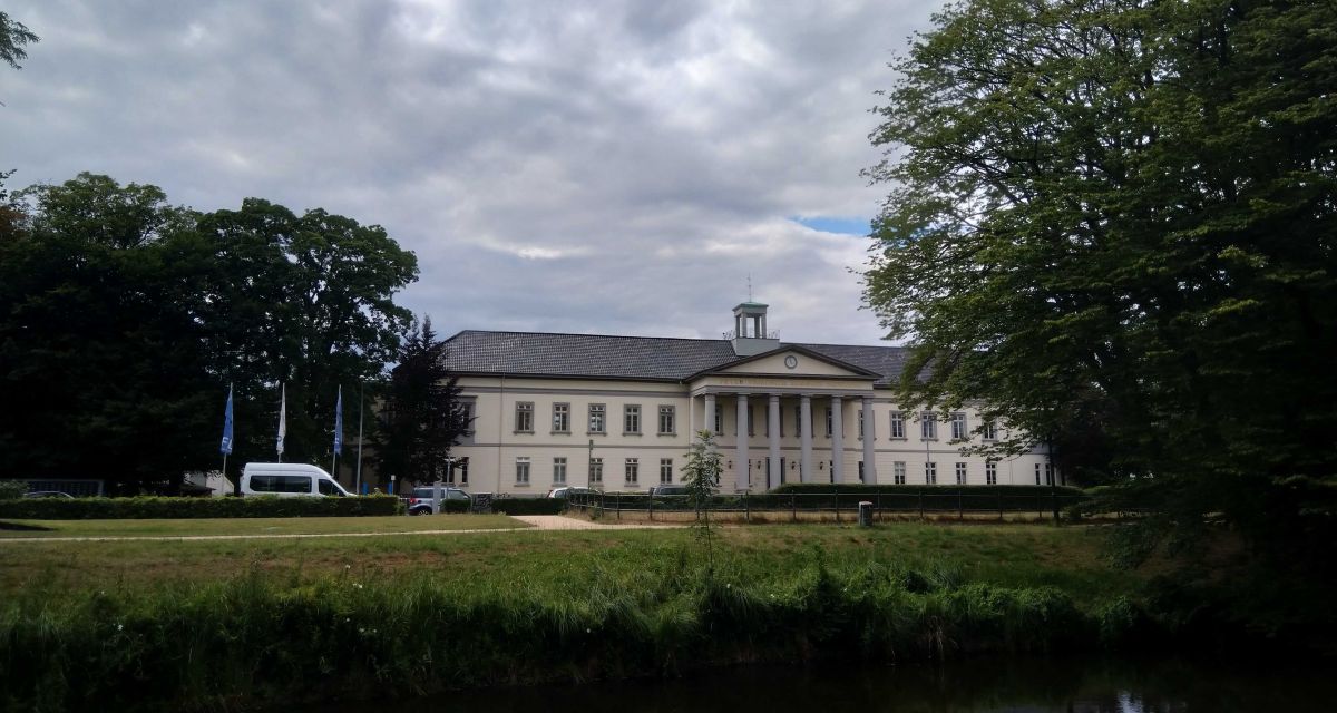 Das Kulturzentrum PFL in Oldenburg. Im ehemaligen Peter-Friedrich-Ludwig-Hospital ist inzwischen ein Kulturzentrum untergebracht, auch die Ratssitzungen der Stadt Oldenburg finden hier statt. Der weiße, klassizistische Bau befindet sich in der Innenstadt, im Vordergrund ist der Fluss Haaren zu sehen, das Gebäude ist auf dem Foto von Bäumen gesäumt, der Himmel ist bewölkt.