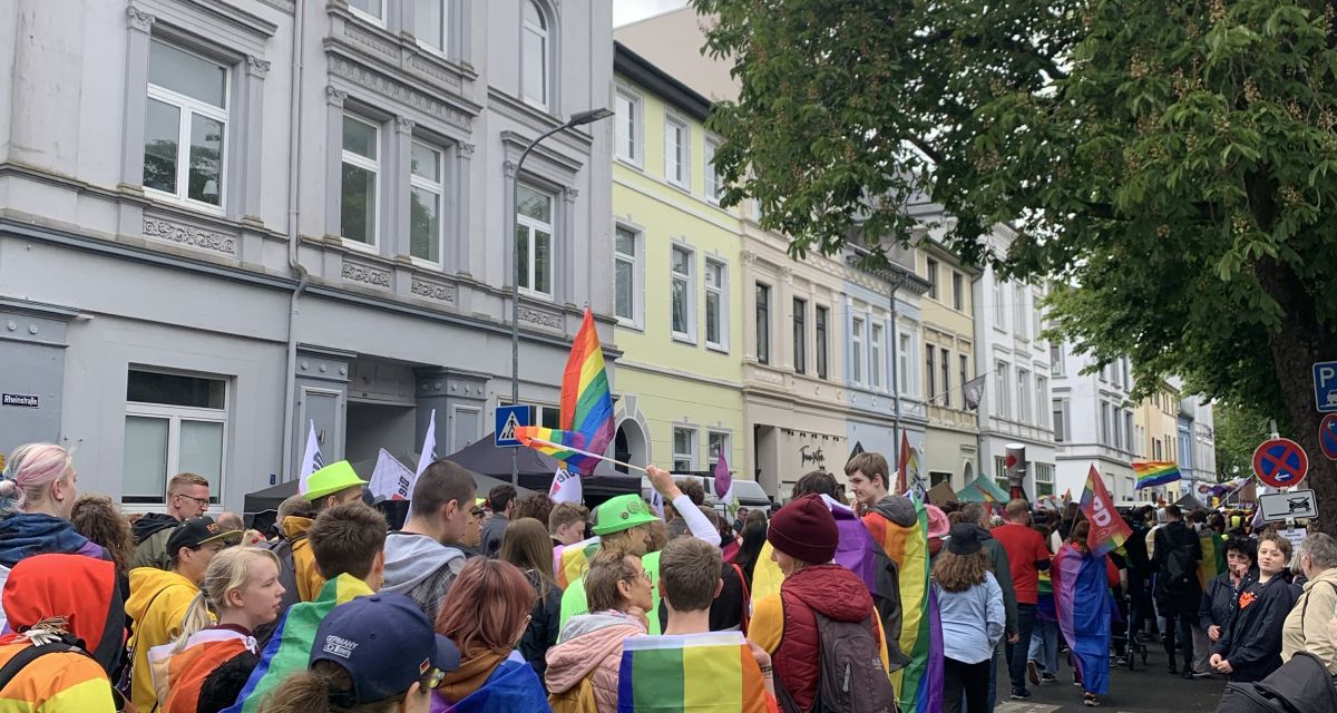 Der CSD Wilhelmshaven zieht durch die Rheinstraße. Viele Menschen sind mit dem Rücken zur Kamera zu sehen, viele tragen Regenbogenflaggen.