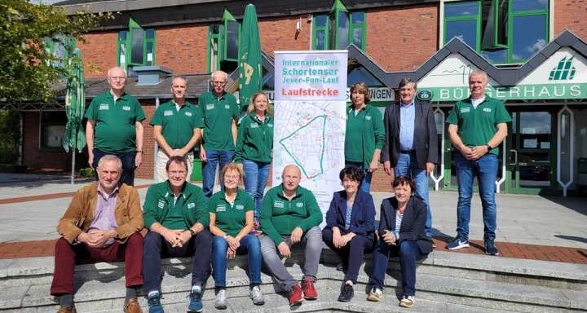 Die Organisator*innen des Jever Fun Lauf in Schortens stehen mit einem großen Aufsteller zur Veranstaltung vor dem Bürgerhaus in Schortens Viele von ihnen tragen grüne Jever-Pilsener-T-Shirts.