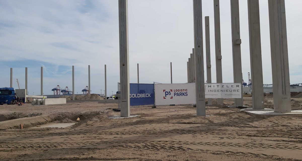 Zu sehen sind die Stützpfeiler der zukünftigen Lagerhallen von P3 Logistic am JadeWeserPort in Wilhelmshaven. Graue Betonpfeiler ragen in die Höhe, der Boden ist sandig. Im Hintergrund sind Transparente der durchführenden Firmen zwischen den Pfeilern zu sehen.