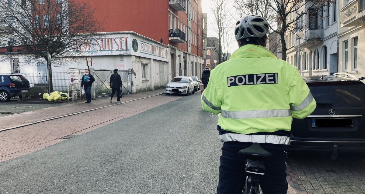 Auf dem Bild ist ein Polizist auf einem Fahrrad zu sehen. Er trägt eine gelbe Warnjacke der Polizei. Nur sein Rücken ist zu sehen. Er trägt einen Helm. 