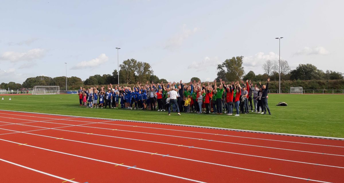 Teilnehmer*innen des Sportfestes zur Eröffnung des Sportparks Varel stehen auf der Rasenfläche und heben die Arme. Im Vordergrund ist die Laufbahn zu sehen.