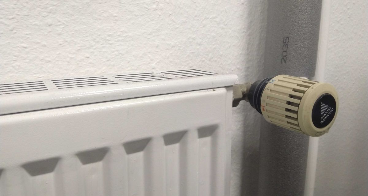 Zu sehen ist ein Heizkörper mit Thermostat, im Hintergrund ein mit Dämmmaterial verkleidetes Heizungsrohr.