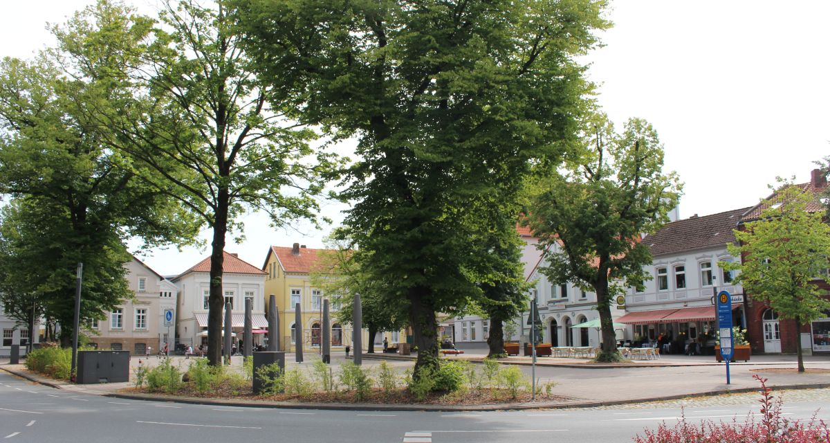 Zu sehen ist der Schlossplatz in Varel an einem Sommertag. Im Vordergrund ist die Straße zu erkennen, im Bildmittelgrund einige Bäume, im Hintergrund historische Gebäude.