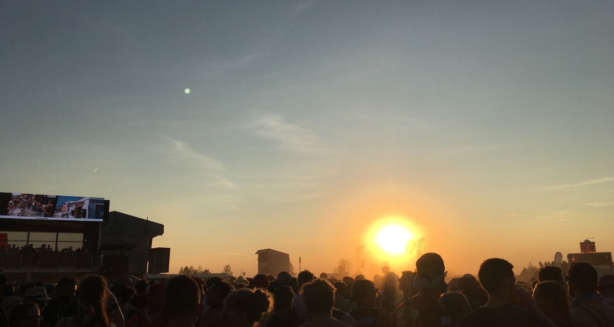 Sonnenuntergang über einem Festival an einem heißen Sommerabend. Im Gegenlicht sind viele Menschenumrisse zu erkennen, außerdem eine Bühne