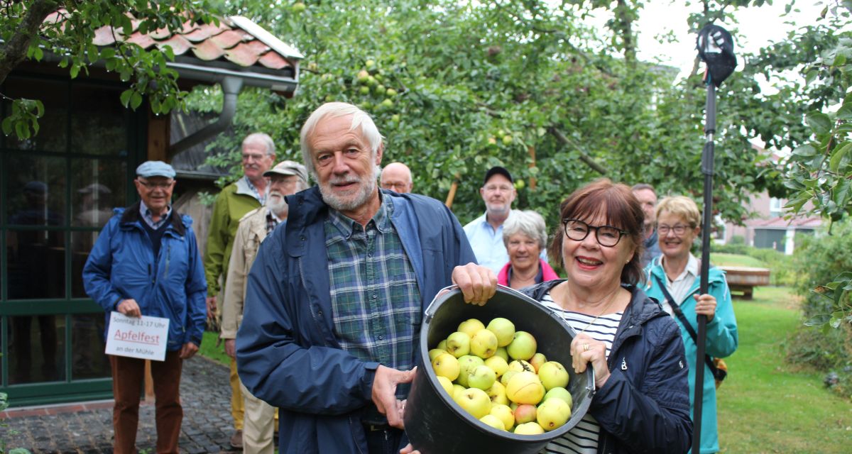 Die Veranstalter*innen stellen das Apfelfest an der Schlachtmühle in Jever vor. Eine große Gruppe steht in einem Apfelgarten, zwei von ihnen halten einen Eimer mit frisch gepflückten Äpfeln in die Kamera.