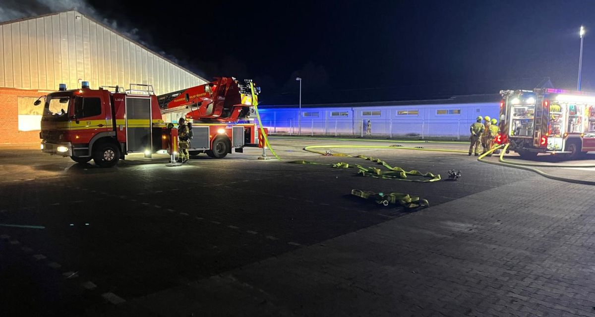 Zu sehen ist das Gebäude eines ehemaligen Discount-Marktes in der Güterstraße in Wilhelmshaven. Aus Fenstern und durch das Dach schlagen Flammen und Rauchsäulen in den dunklen Nachthimmel. Im Vordergrund sind zwei Einsatzfahrzeuge der Feuerwehr Wilhelmshaven zu sehen, davor liegen mehrere Löschschläuche. Feuerwehrleute stehen an den Fahrzeugen.