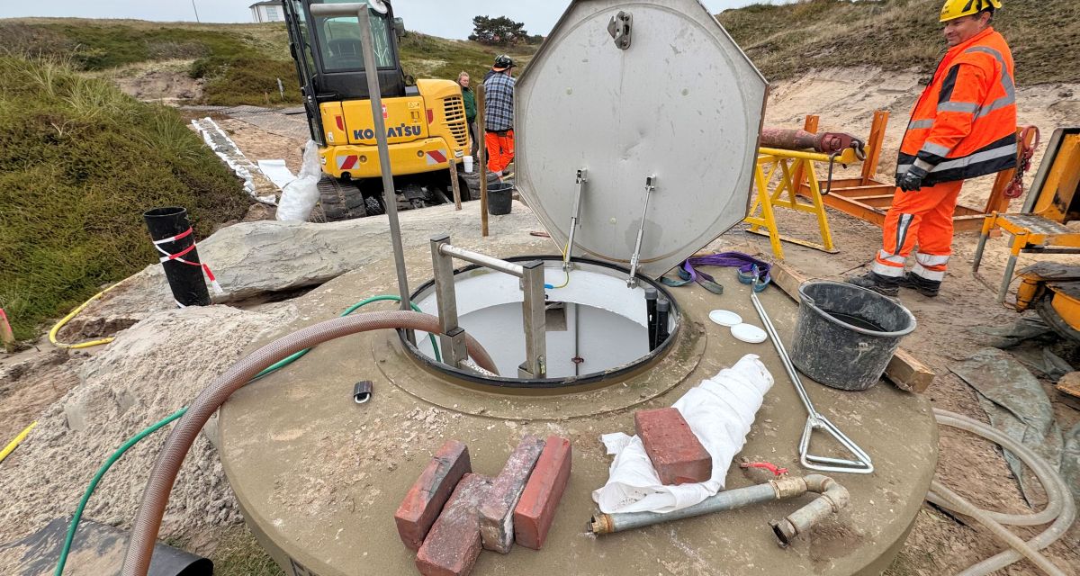 Zu sehen ist einer der sechs Förderbrunnen auf Spiekeroog, die Wasser aus der Süßwasserlinse der Insel an die Oberflüche befördern. Die achteckige Brunnenluke ist geöffnet, ein dicker Schlauch ist hineingeleitet. Um den Brunnen herum sind Baumaterialien, Werkzeug, drei Menschen in Arbeitskleidung und ein Baufahrzeug zu erkennen.